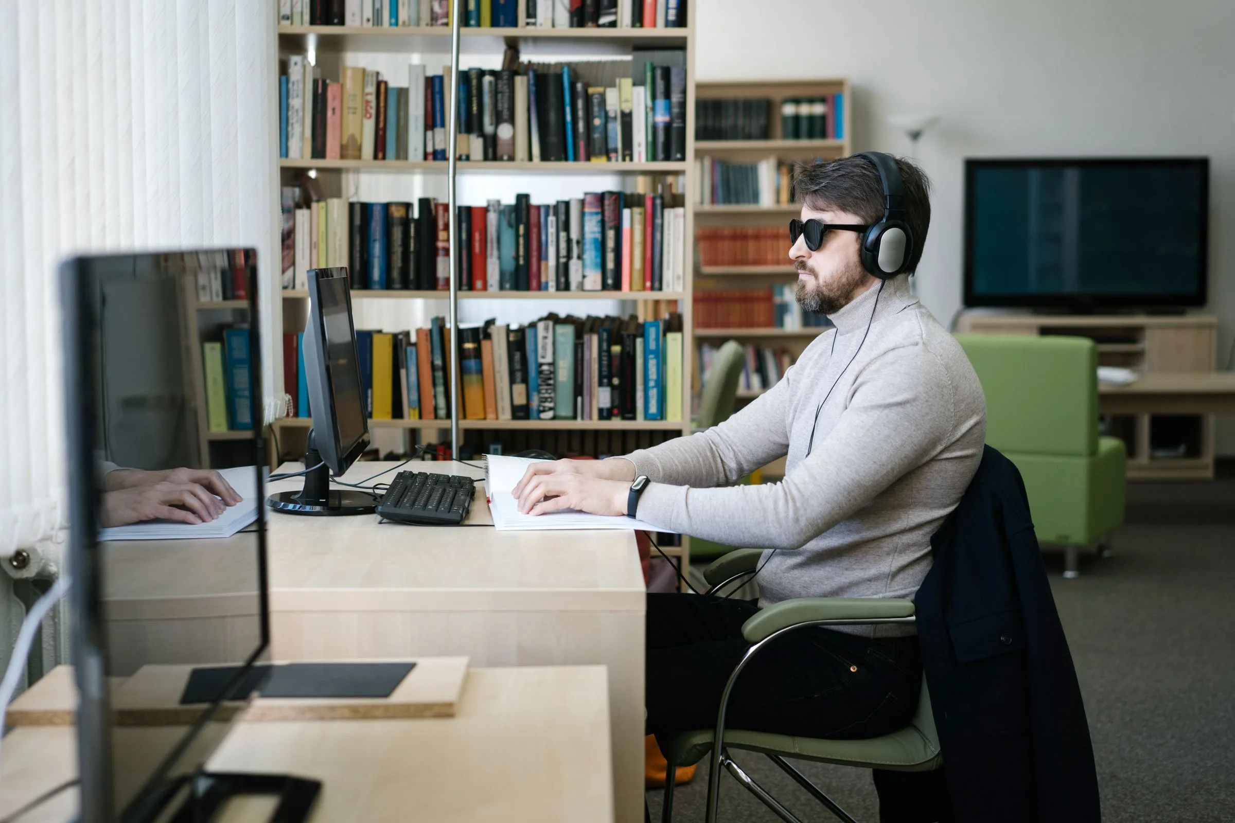 A blind man in headphones and dark glasses works at a desktop computer in a library