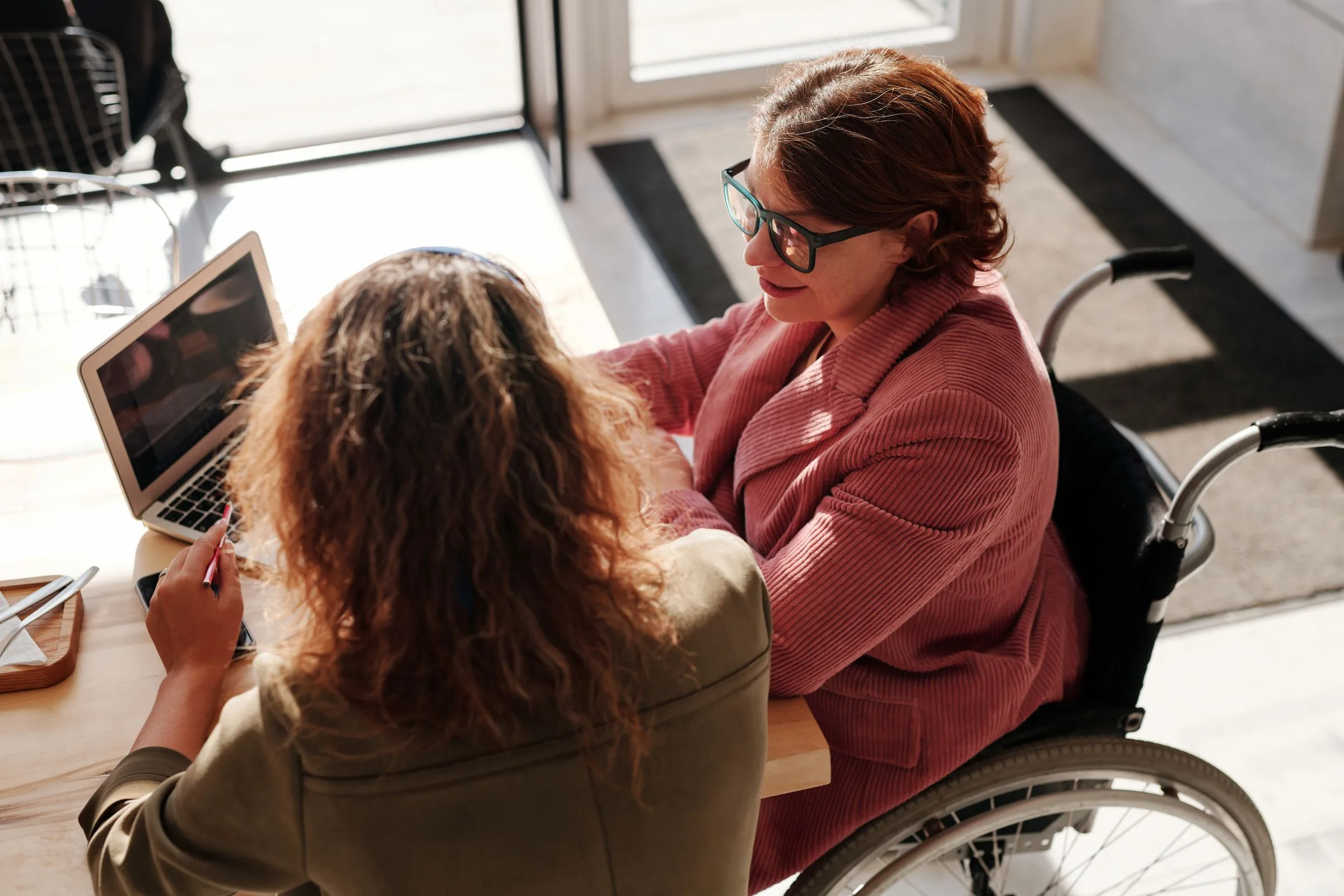 Two women working on laptops at a table by the window, one of them a wheelchair user