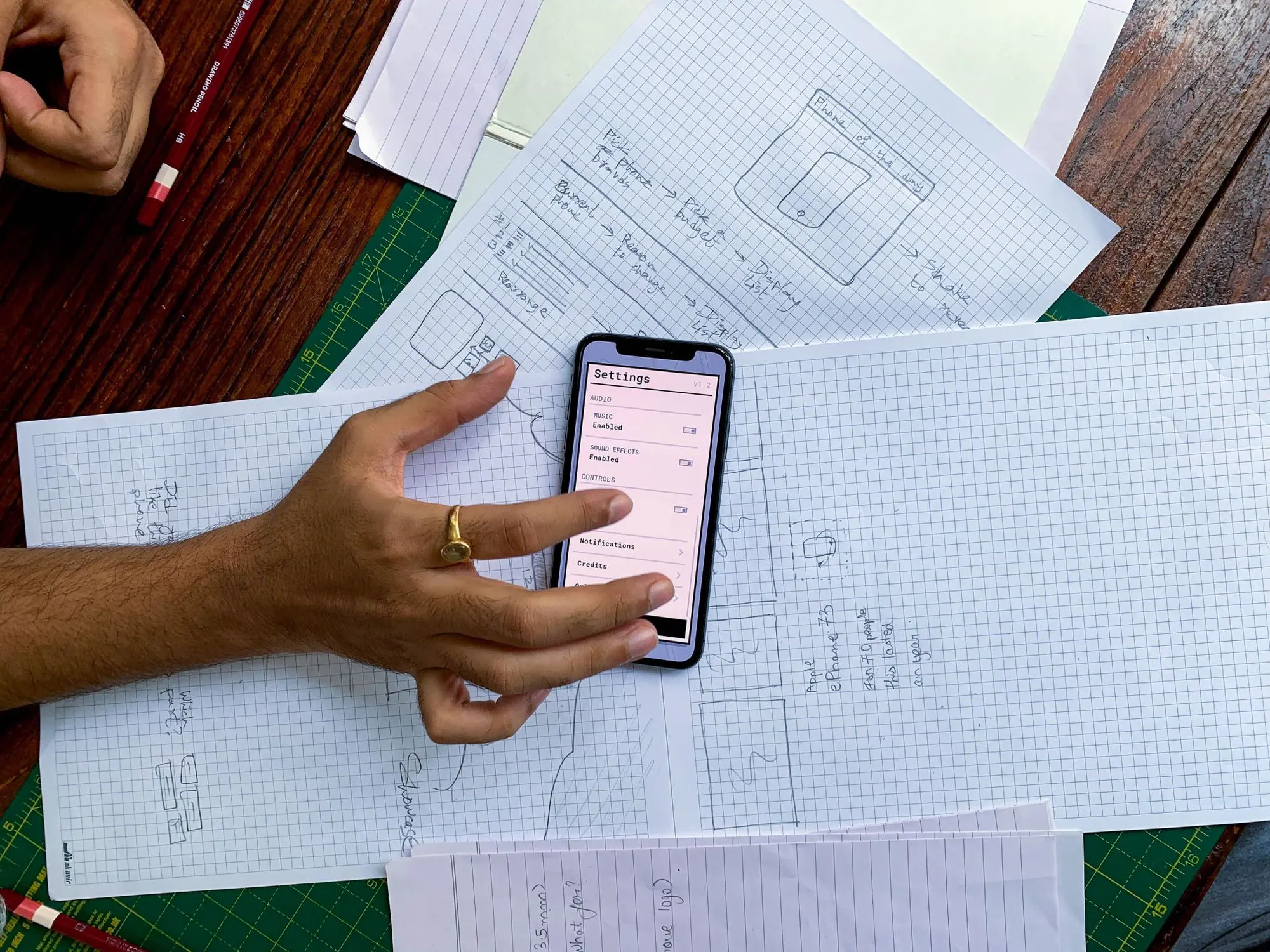 A hand holds a phone running an app prototype over the paper wireframe sketches it was designed from